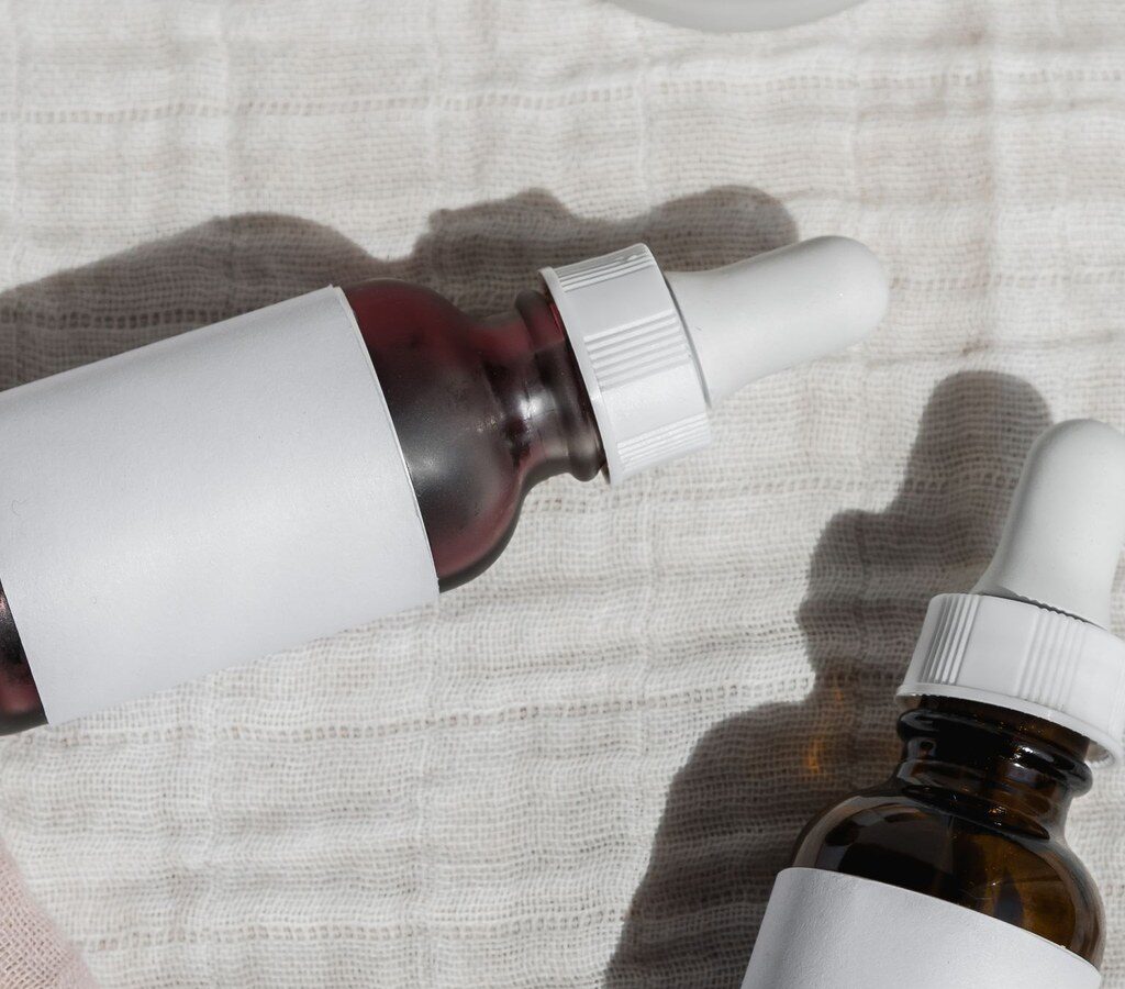 Zwei Kosmetikflaschen mit Pipette und neutralen weißen Etiketten auf hellem Stoff