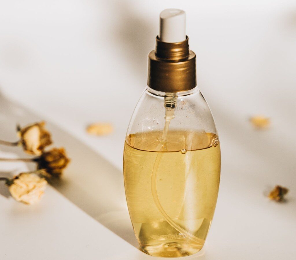 Gläserne Sprühflasche mit gelblicher Flüssigkeit, umgeben von getrockneten Blüten auf hellem Untergrund