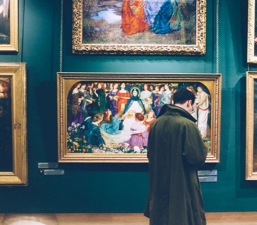 Eine Person steht von hinten vor mehreren gerahmten Gemälden an einer grünen Galeriewand