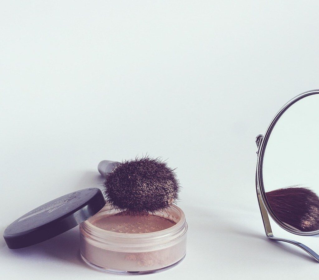 Loser Puder in einer offenen Dose mit Pinsel neben einem kleinen Standspiegel auf hellem Hintergrund