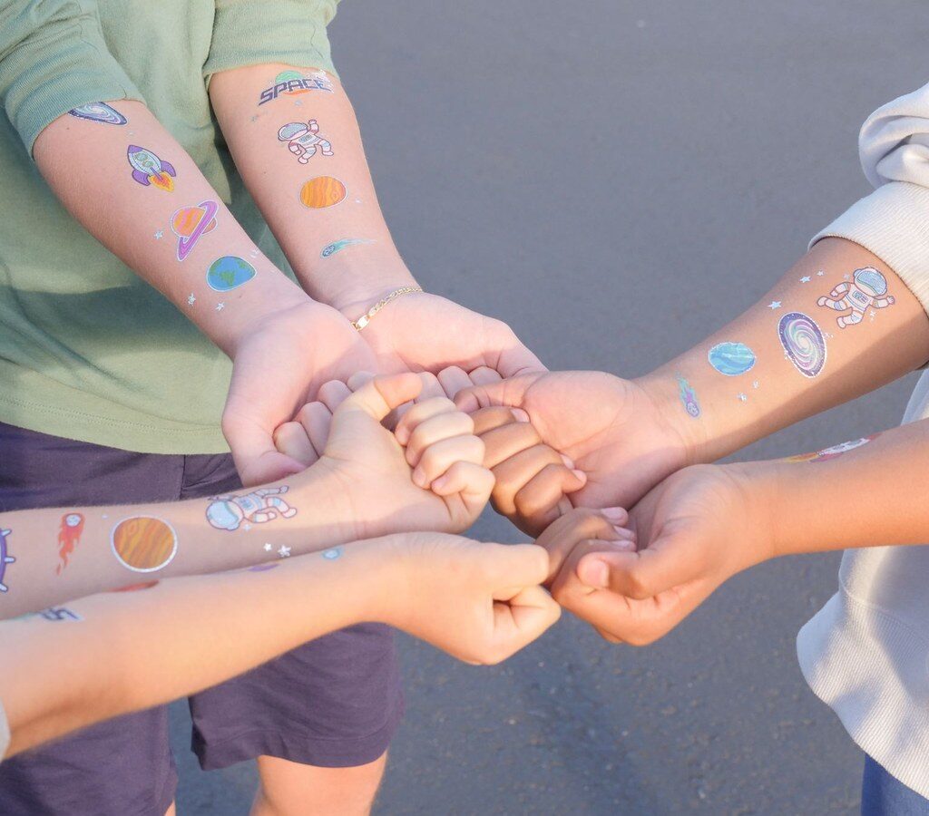 Vier Kinderarme mit bunten Klebe-Tattoos in Weltraummotiven, die Hände sind in der Mitte zusammengelegt